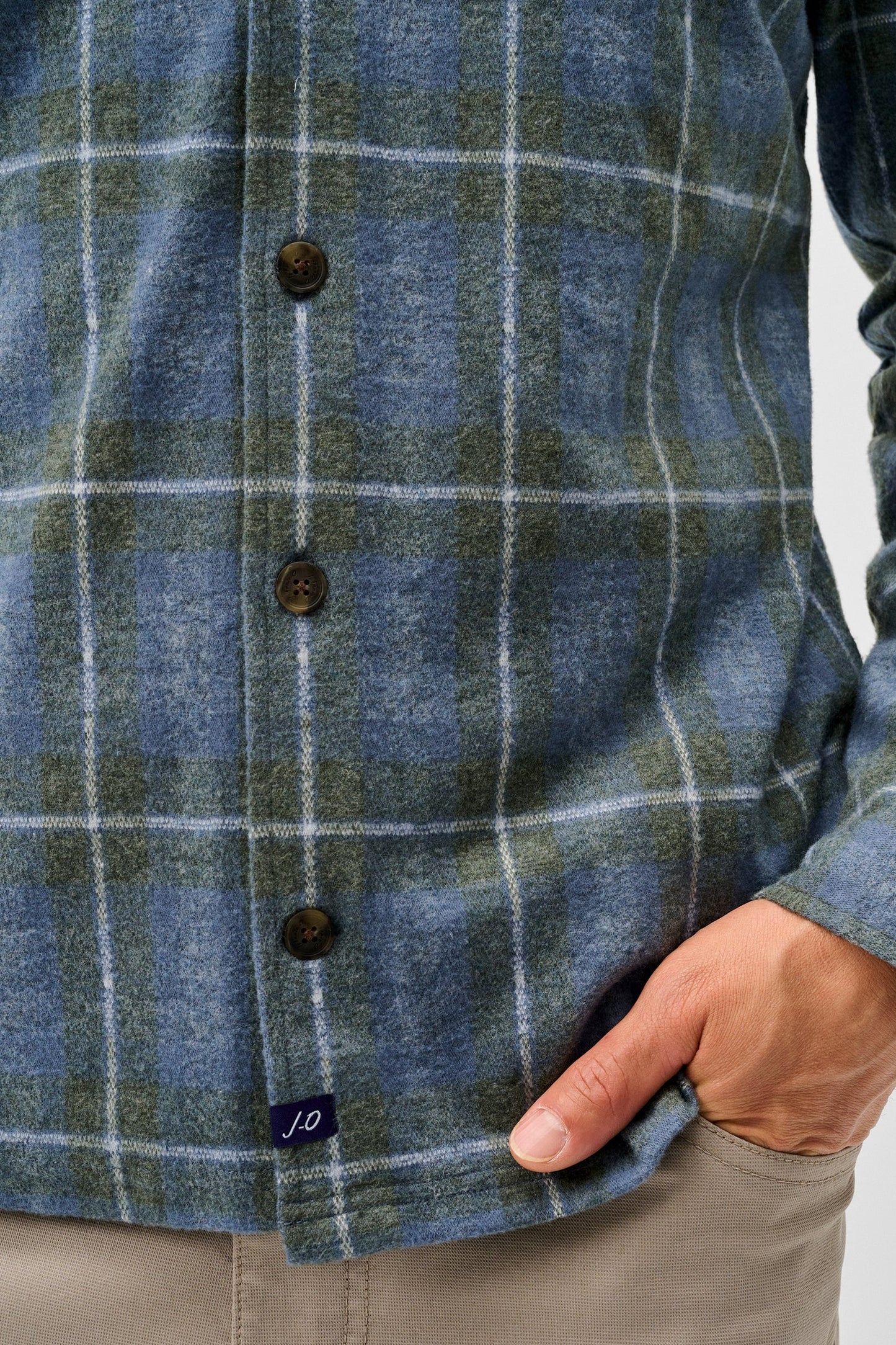 Close-up of a person wearing a blue and green plaid button-up shirt with brown buttons, paired with beige trousers, against a light gray background.