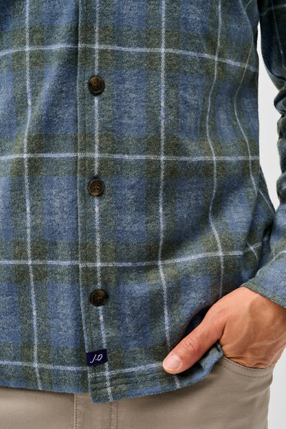 Close-up of a person wearing a blue and green plaid button-up shirt with brown buttons, paired with beige trousers, against a light gray background.