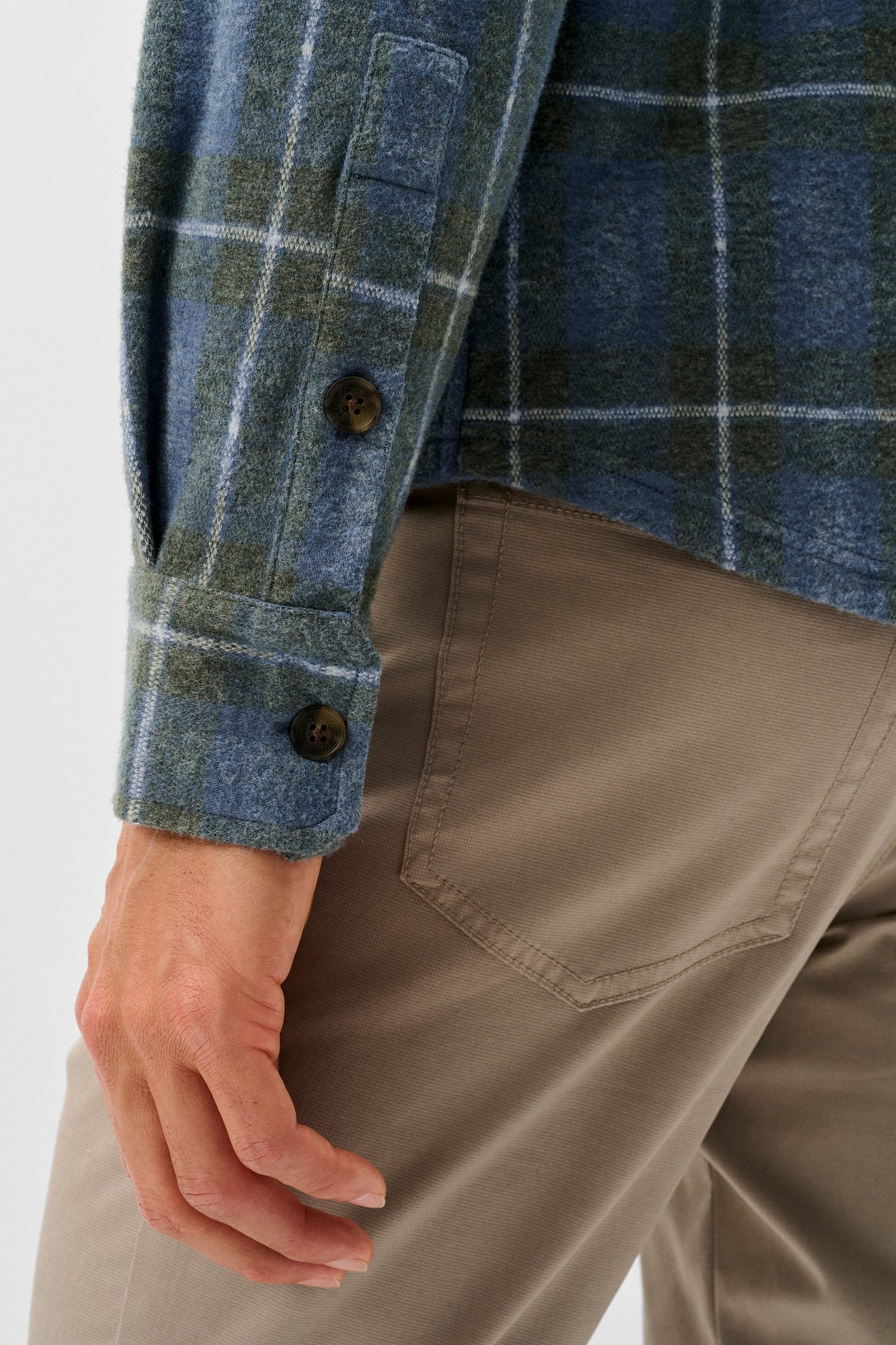 Close-up of a person wearing a blue and green plaid flannel shirt with brown buttons and tan trousers, showing the sleeve cuff and side pocket area.