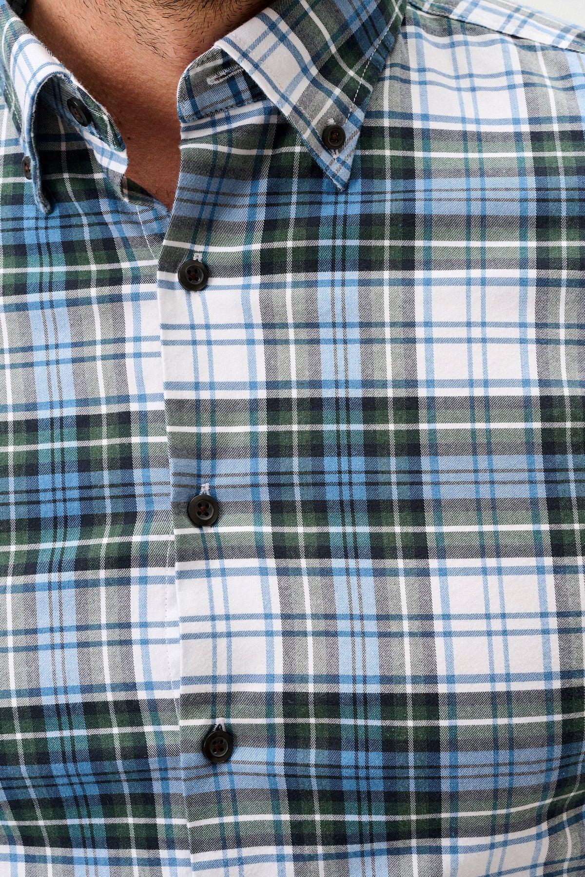 Close-up of a men's button-down plaid shirt worn by a model, featuring a pattern of white, light blue, dark green, and gray intersecting plaid stripes.