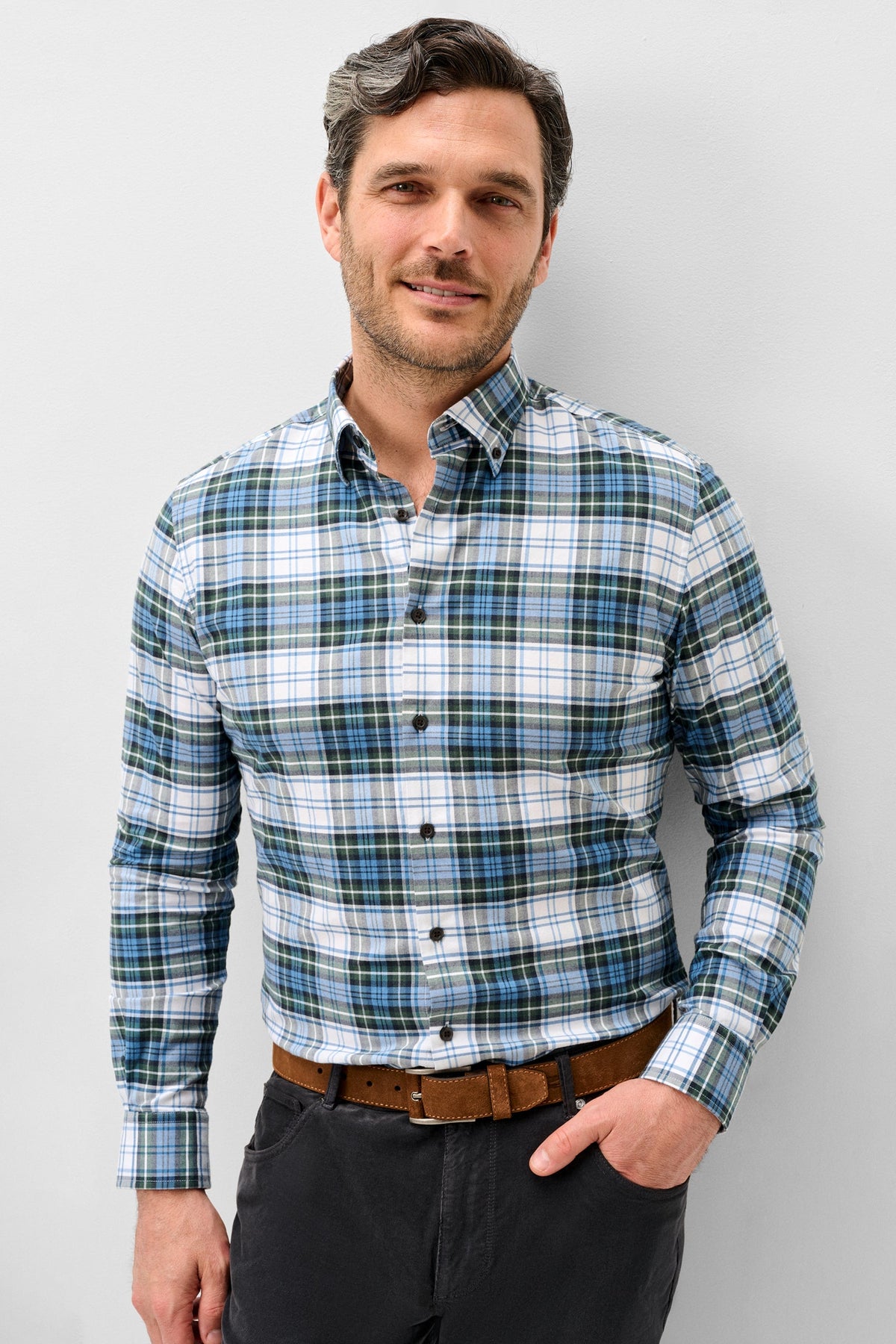 A man wearing a long-sleeve plaid button-down shirt in blue, green, and white, paired with a brown belt and dark gray trousers, standing against a light gray background.
