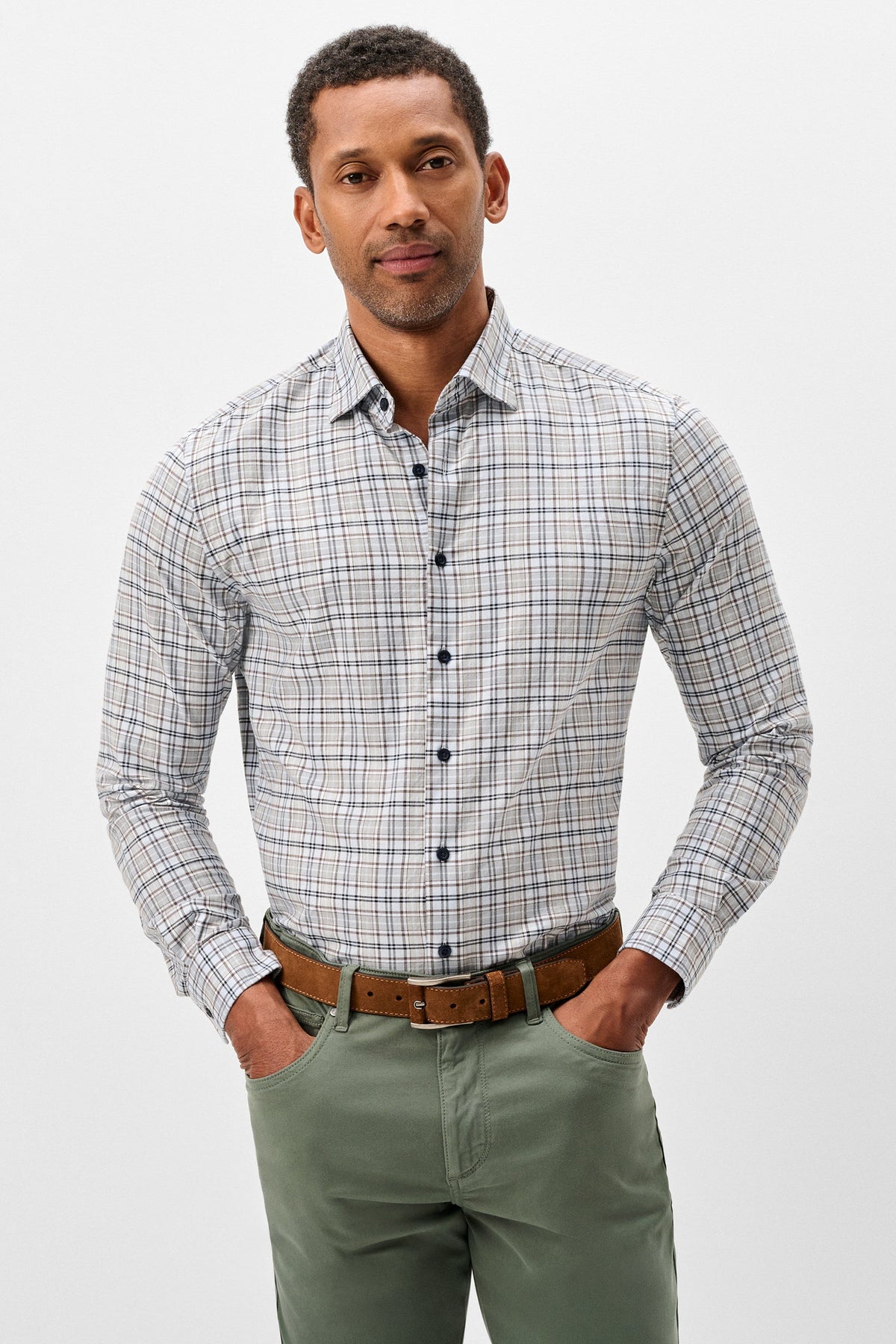 A man wearing a long-sleeve button-down plaid shirt in gray, white, and blue, paired with olive green pants and a brown leather belt, standing against a light gray studio background.