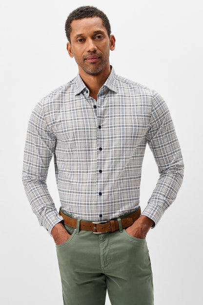 A man wearing a long-sleeve button-down plaid shirt in gray, white, and blue, paired with olive green pants and a brown leather belt, standing against a light gray studio background.