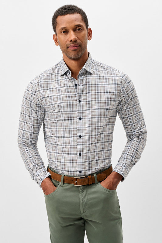 A man wearing a long-sleeve button-down plaid shirt in gray, white, and blue, paired with olive green pants and a brown leather belt, standing against a light gray studio background.