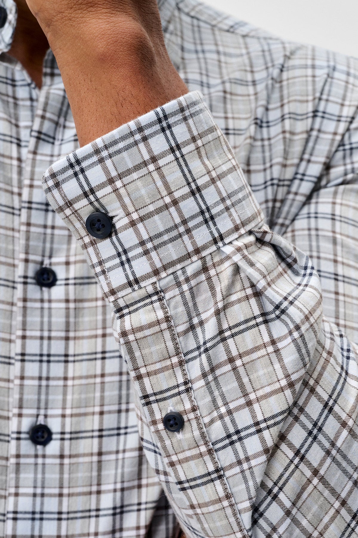 Close-up of a person wearing a long-sleeve plaid button-up shirt, showing the cuff, buttons, and part of the torso against a light neutral background.