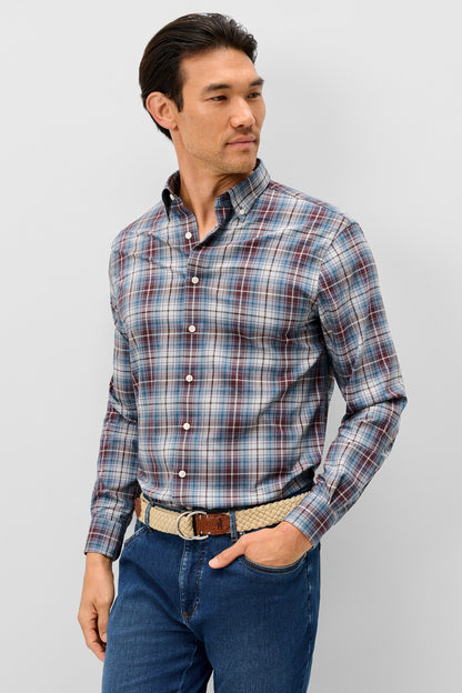 A man wearing a long-sleeve button-down plaid shirt in blue, red, and white, paired with blue jeans and a patterned belt, standing against a light gray studio background.