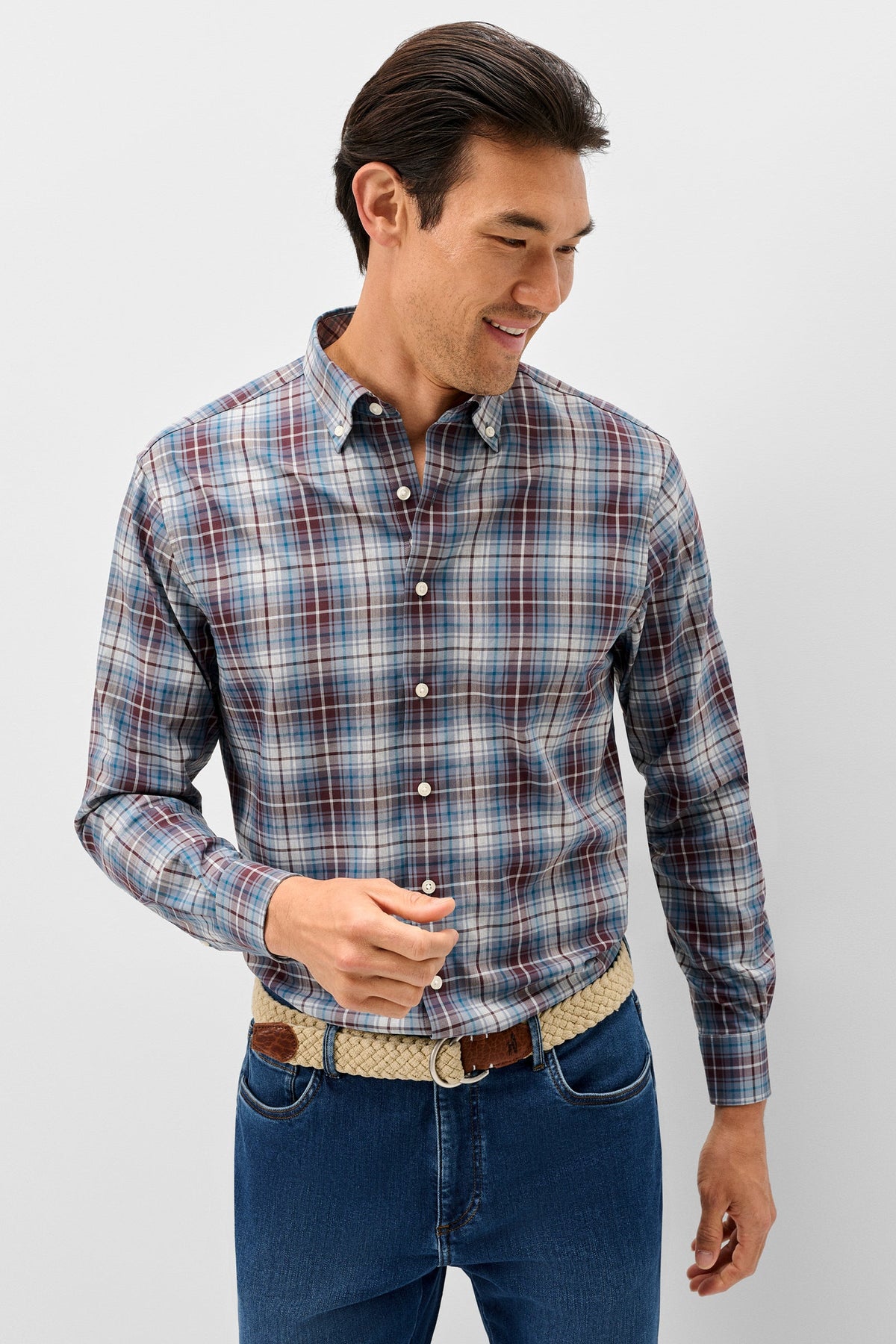 A man wearing a long-sleeve plaid button-down shirt in blue, red, and white, paired with blue jeans and a patterned belt, standing against a light gray background.