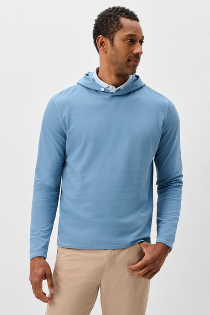 A man wearing a light blue long-sleeve hooded shirt over a white collared shirt, paired with beige pants, standing against a plain light gray studio background.