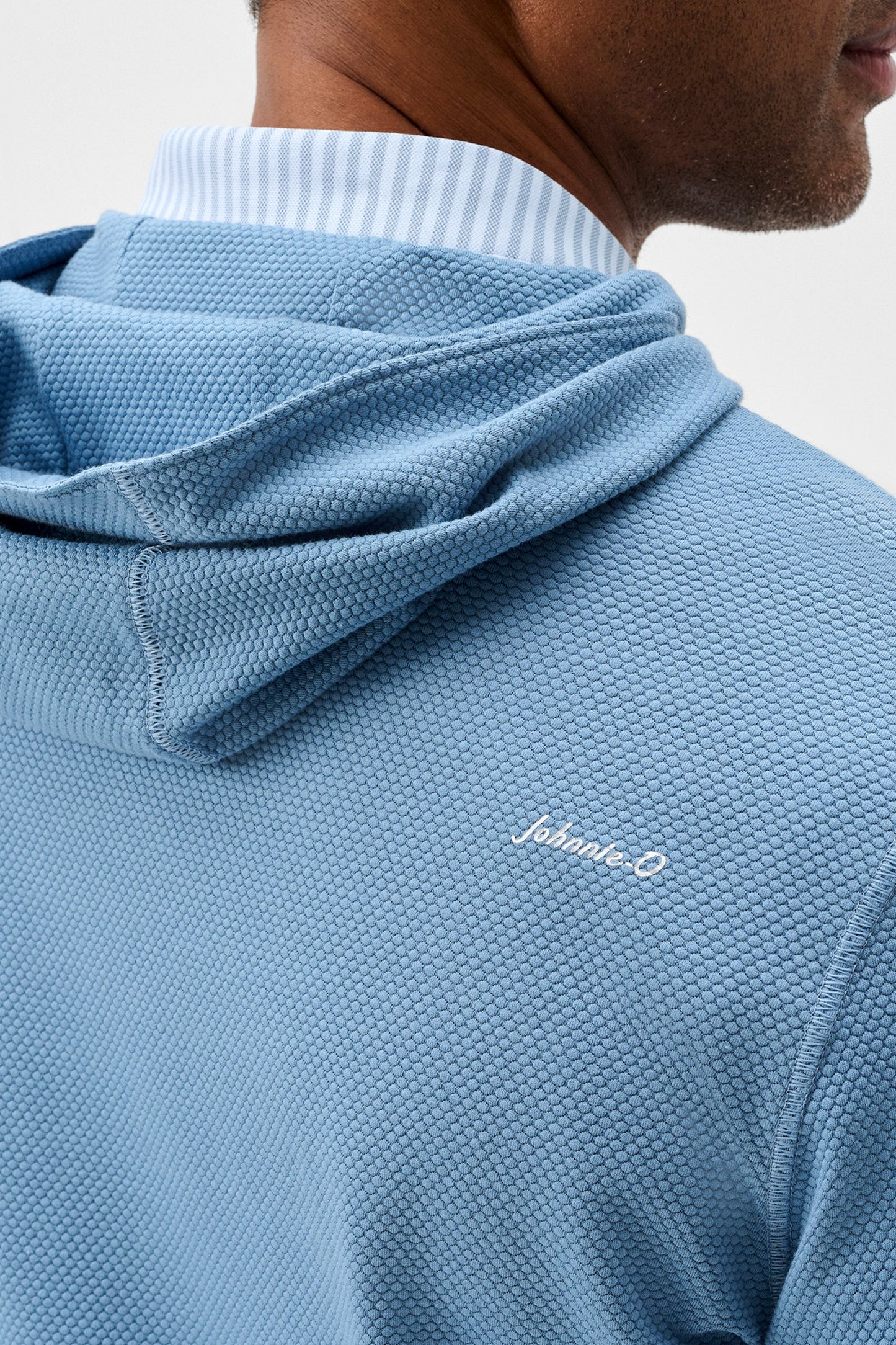 Close-up of a man wearing a light blue textured hooded sweatshirt over a white and blue striped collared shirt, with a small white script logo on the chest.
