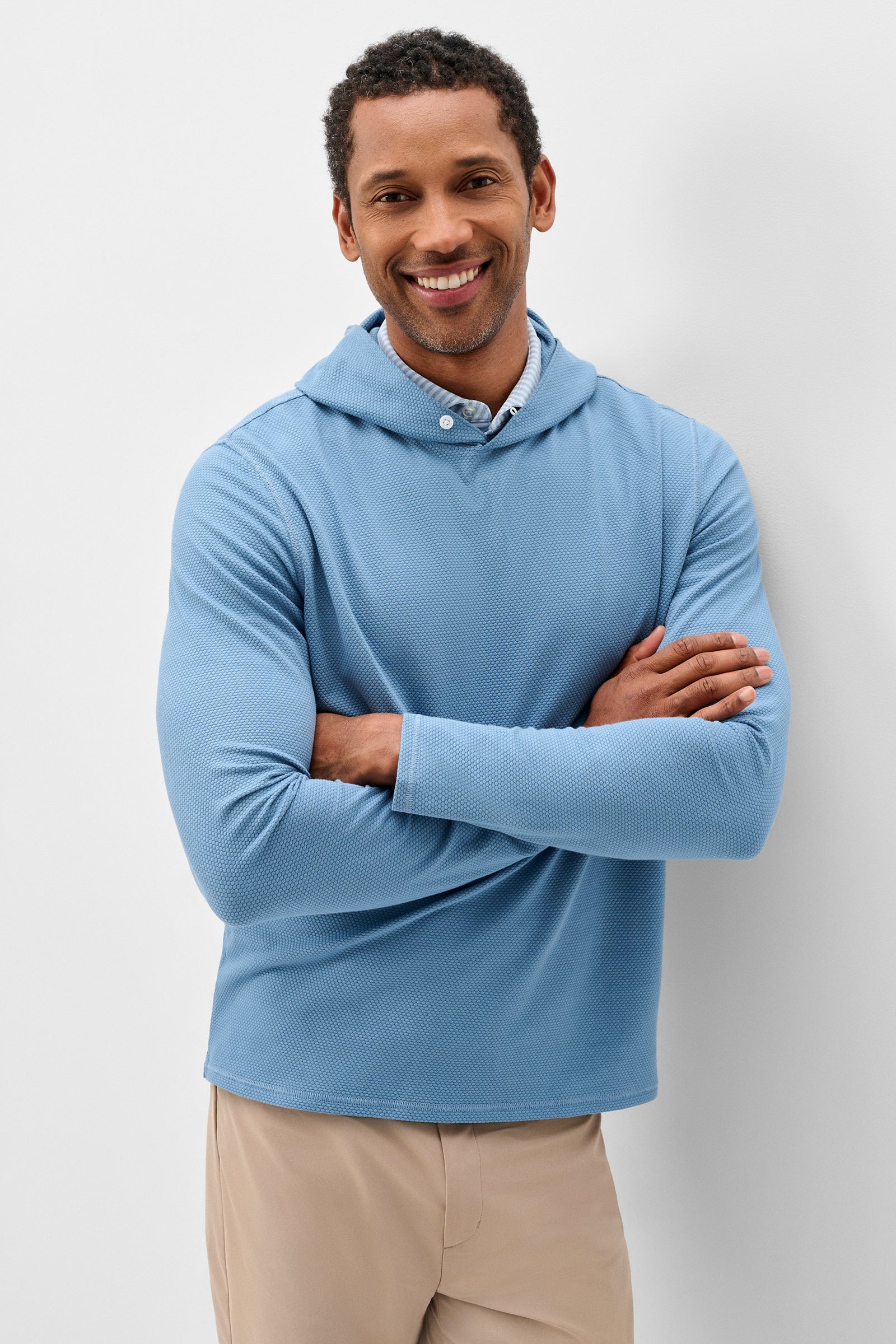 A smiling man wearing a light blue long-sleeve hooded shirt with a buttoned collar, paired with beige pants, standing against a light gray studio background.