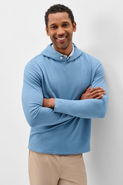 A smiling man wearing a light blue long-sleeve hooded shirt with a buttoned collar, paired with beige pants, standing against a light gray studio background.