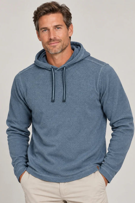 A man wearing a heathered slate blue long-sleeve hoodie with dark drawstrings, paired with light beige trousers, standing against a neutral light gray background.