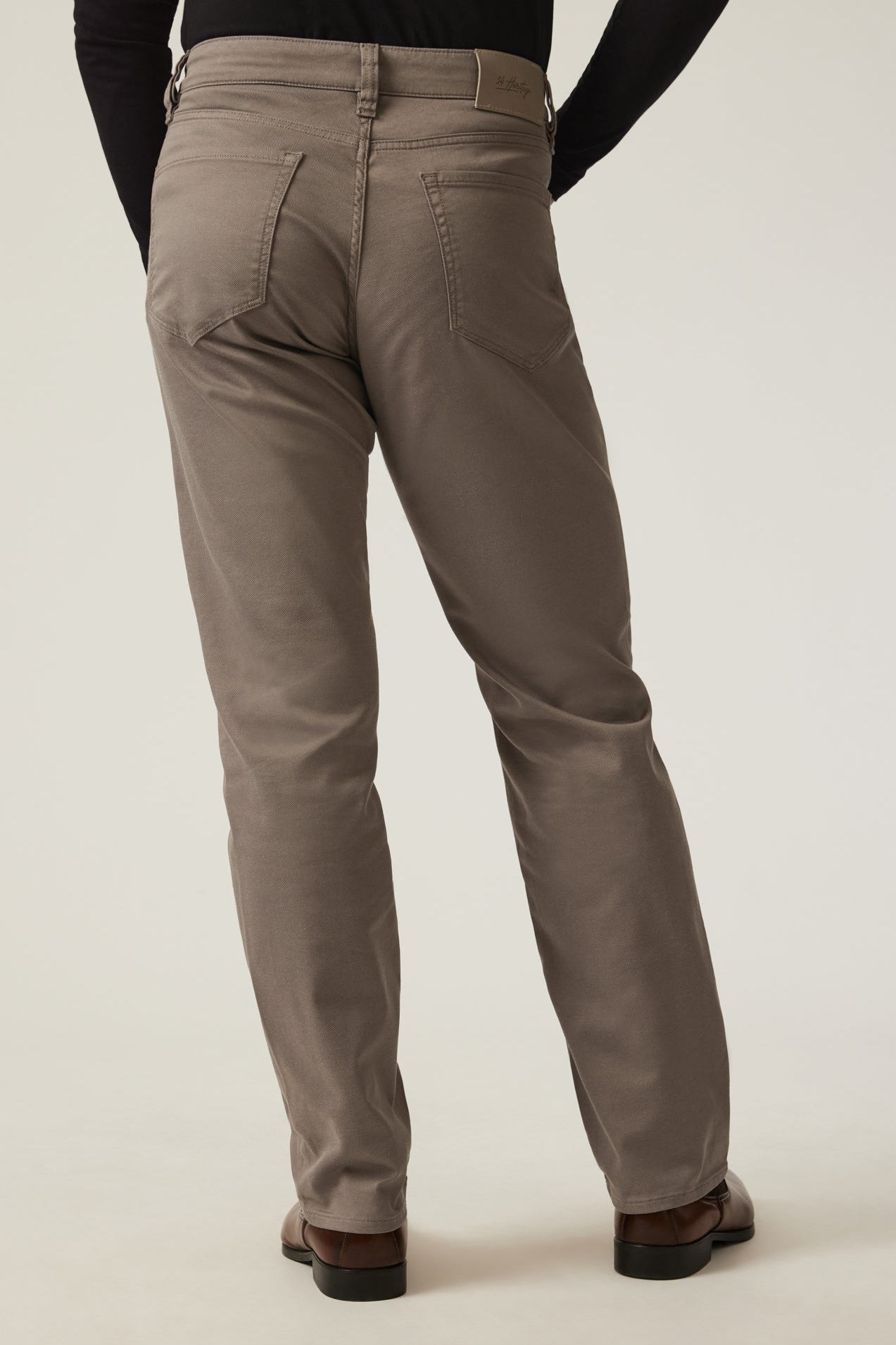 Rear view of a person wearing taupe-brown straight-leg chinos with a black long-sleeve top and brown leather shoes, against a light beige studio background.