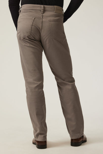 Rear view of a person wearing taupe-brown straight-leg chinos with a black long-sleeve top and brown leather shoes, against a light beige studio background.