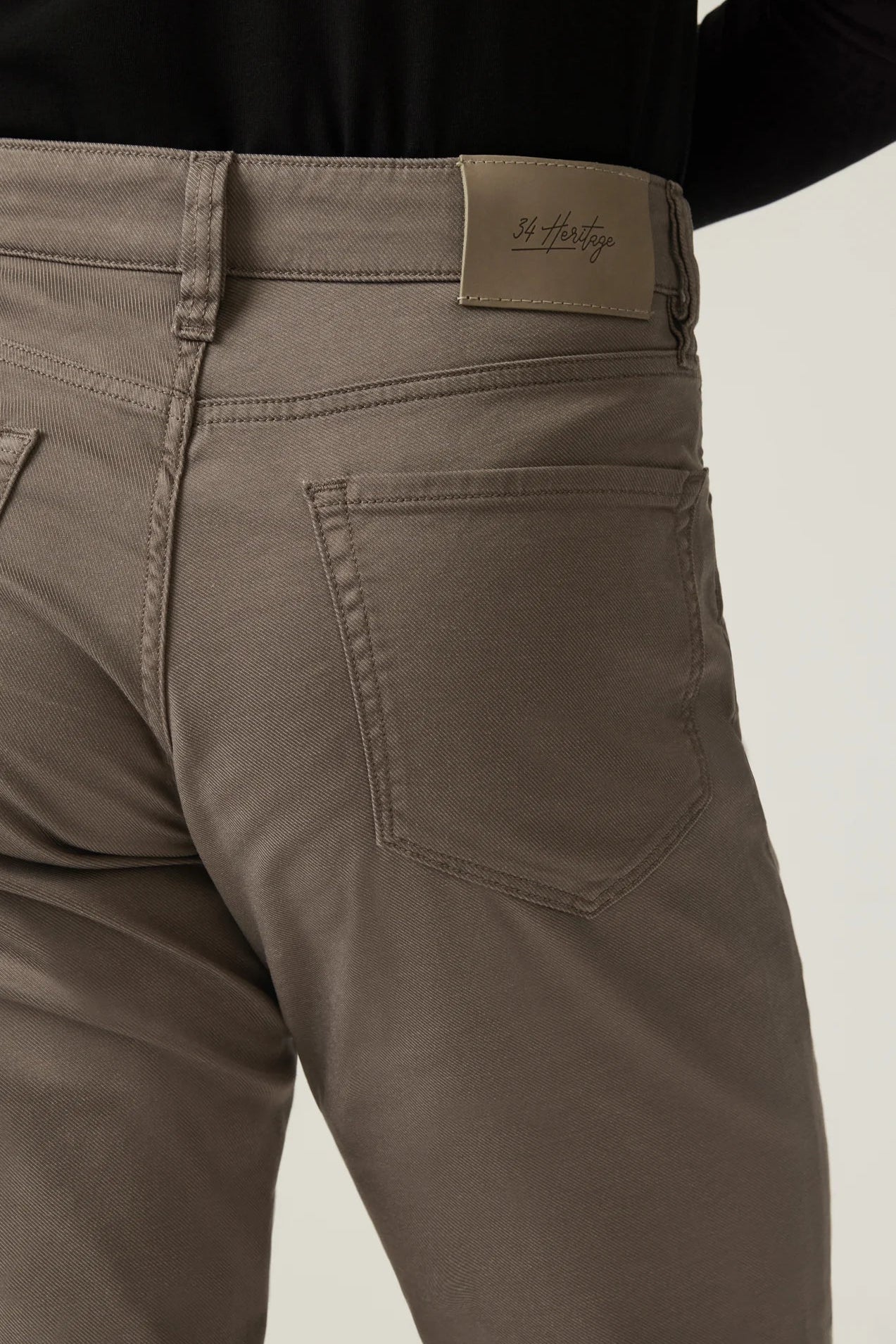 Close-up of the back waistband and rear pocket area of a pair of taupe-brown jeans on a model, showing a St. Herberts brand patch on the waistband.