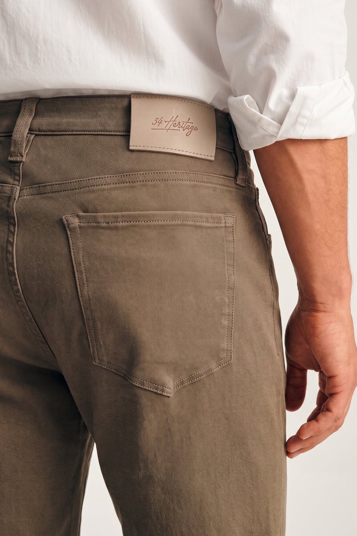 Close-up of a person wearing olive brown denim jeans with a white shirt, showing the back pocket and a leather patch with the brand name '36 Hartley'.