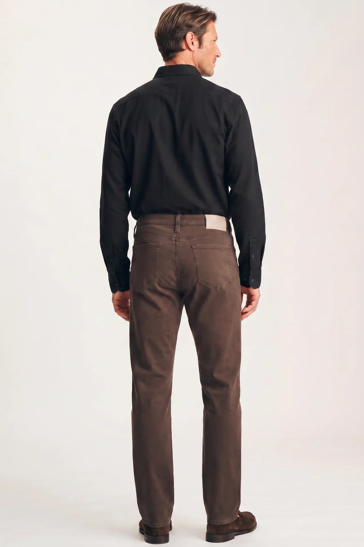 A man viewed from the back wearing a black long-sleeve button-up shirt and brown trousers, standing against a plain light studio background.
