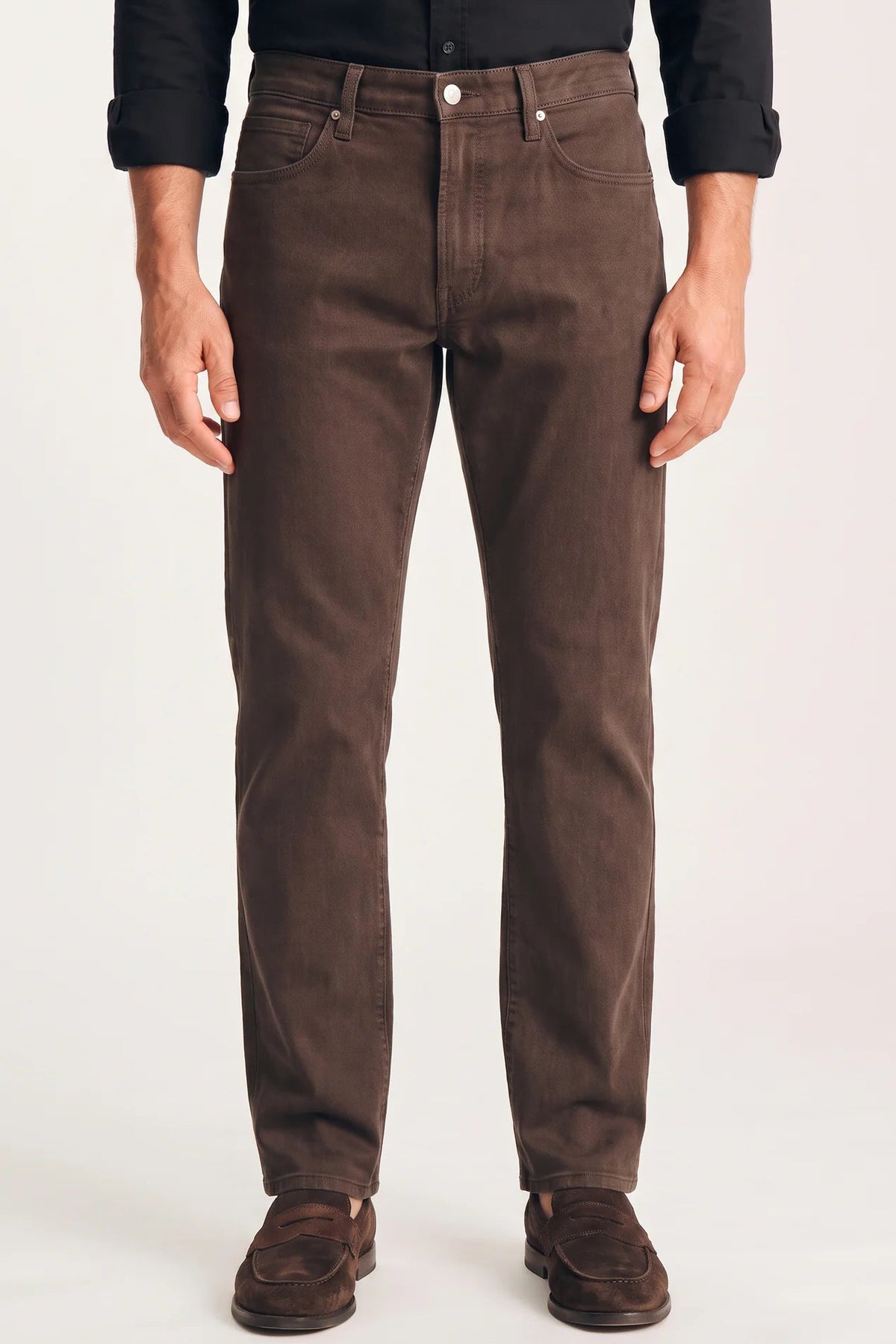 A person wearing dark brown slim-fit jeans, paired with a black long-sleeve shirt and brown suede loafers, standing against a light gray studio background.