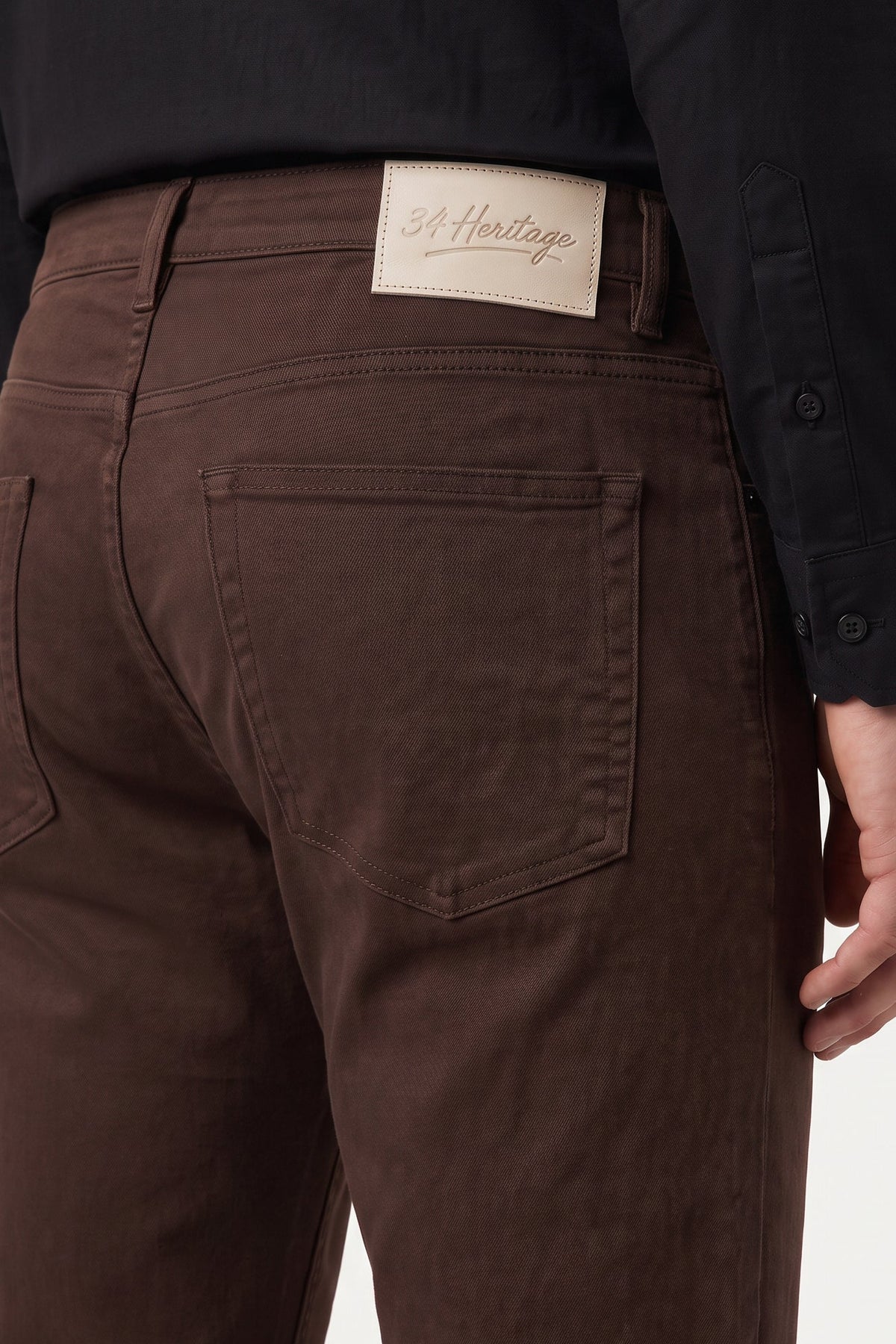 Close-up back view of a person wearing dark brown jeans with a visible St. Heritage brand patch on the waistband, paired with a black shirt.