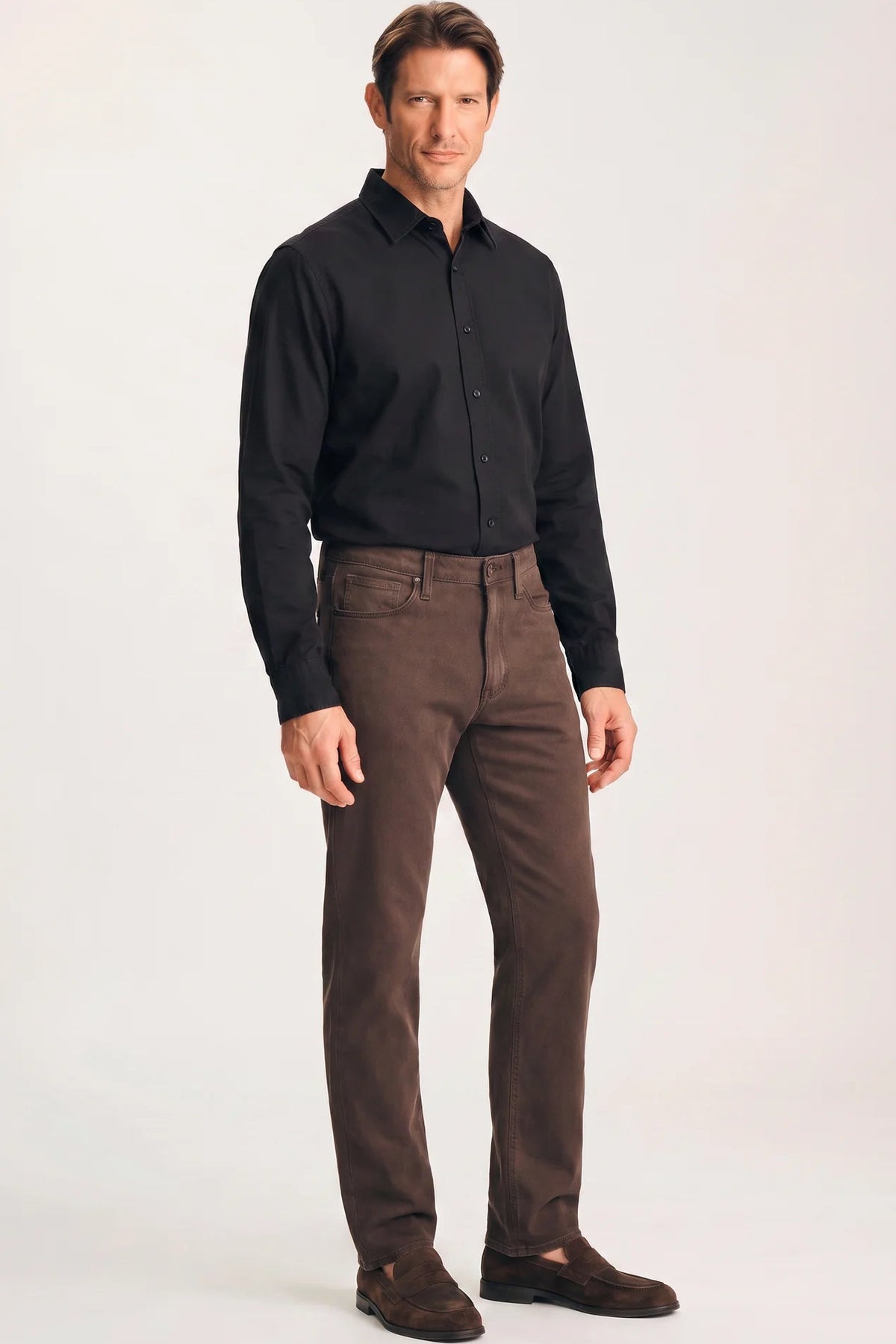 A man modeling a black long-sleeve button-down shirt and brown trousers against a plain light gray studio background.