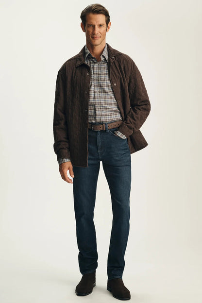 A man modeling a dark brown quilted jacket over a plaid button-down shirt, paired with dark blue jeans and brown suede boots, against a plain off-white studio background.