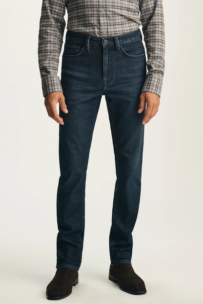 A person wearing dark blue slim-fit jeans, a brown and beige plaid button-down shirt, and dark brown suede boots, standing against a light gray studio background.