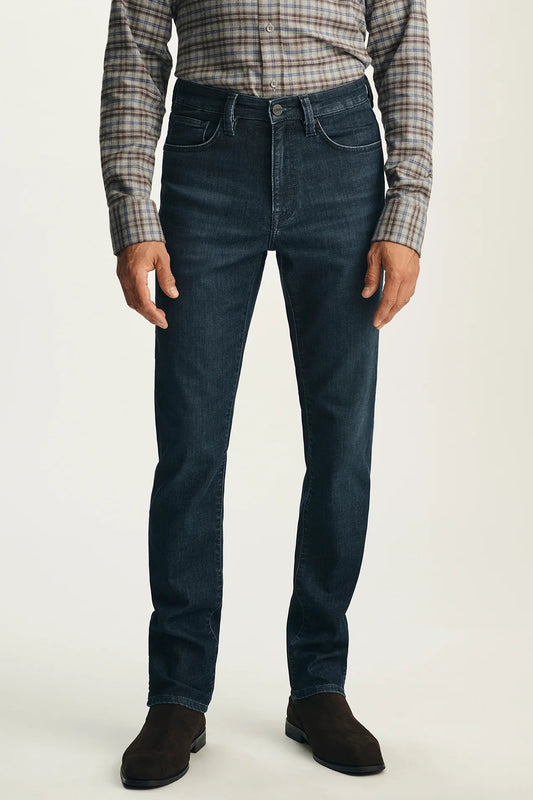 A person wearing dark blue slim-fit jeans, a brown and beige plaid button-down shirt, and dark brown suede boots, standing against a light gray studio background.