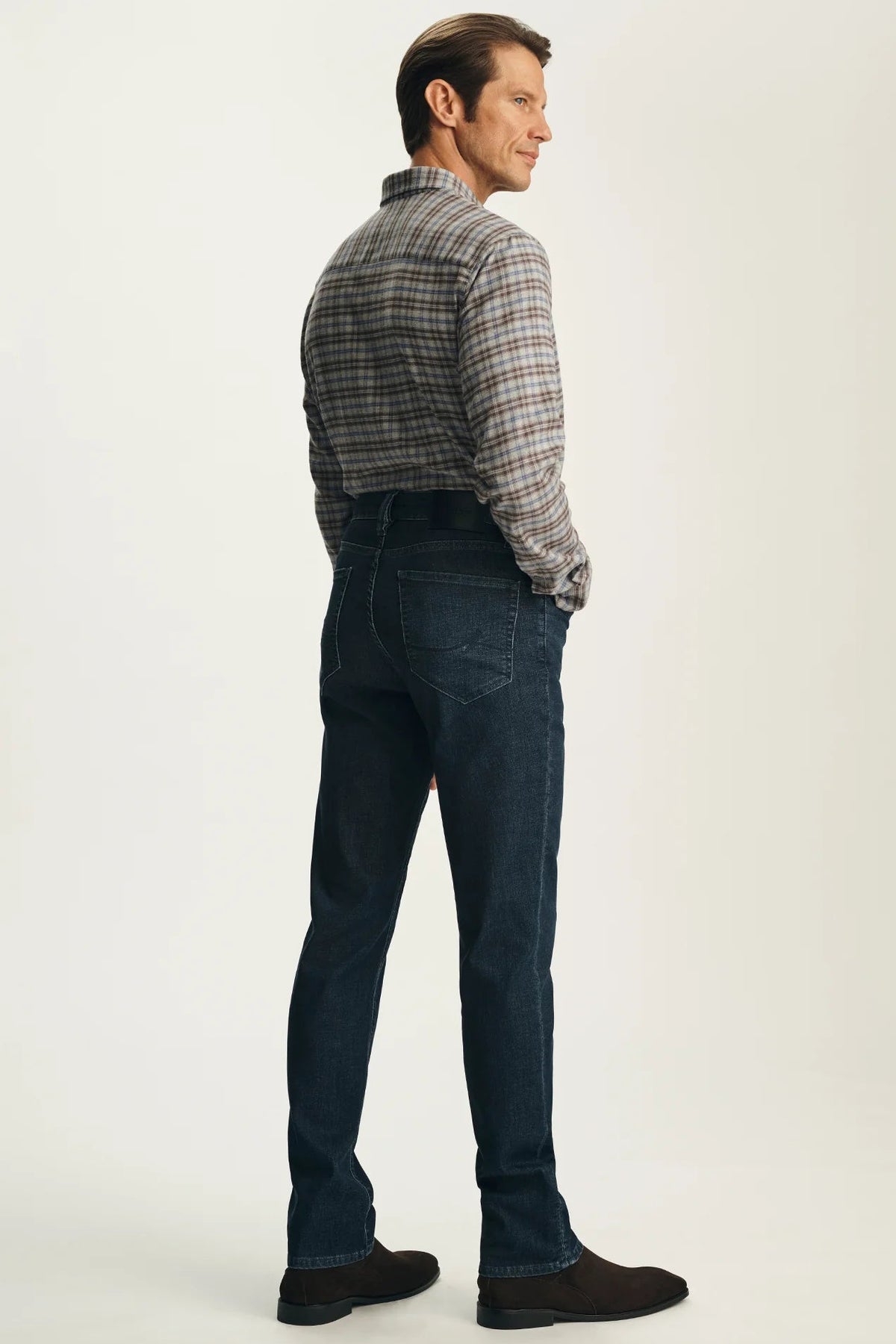 A man shown from the back wearing a gray and brown plaid button-down shirt and dark blue jeans, standing against a plain off-white studio background.