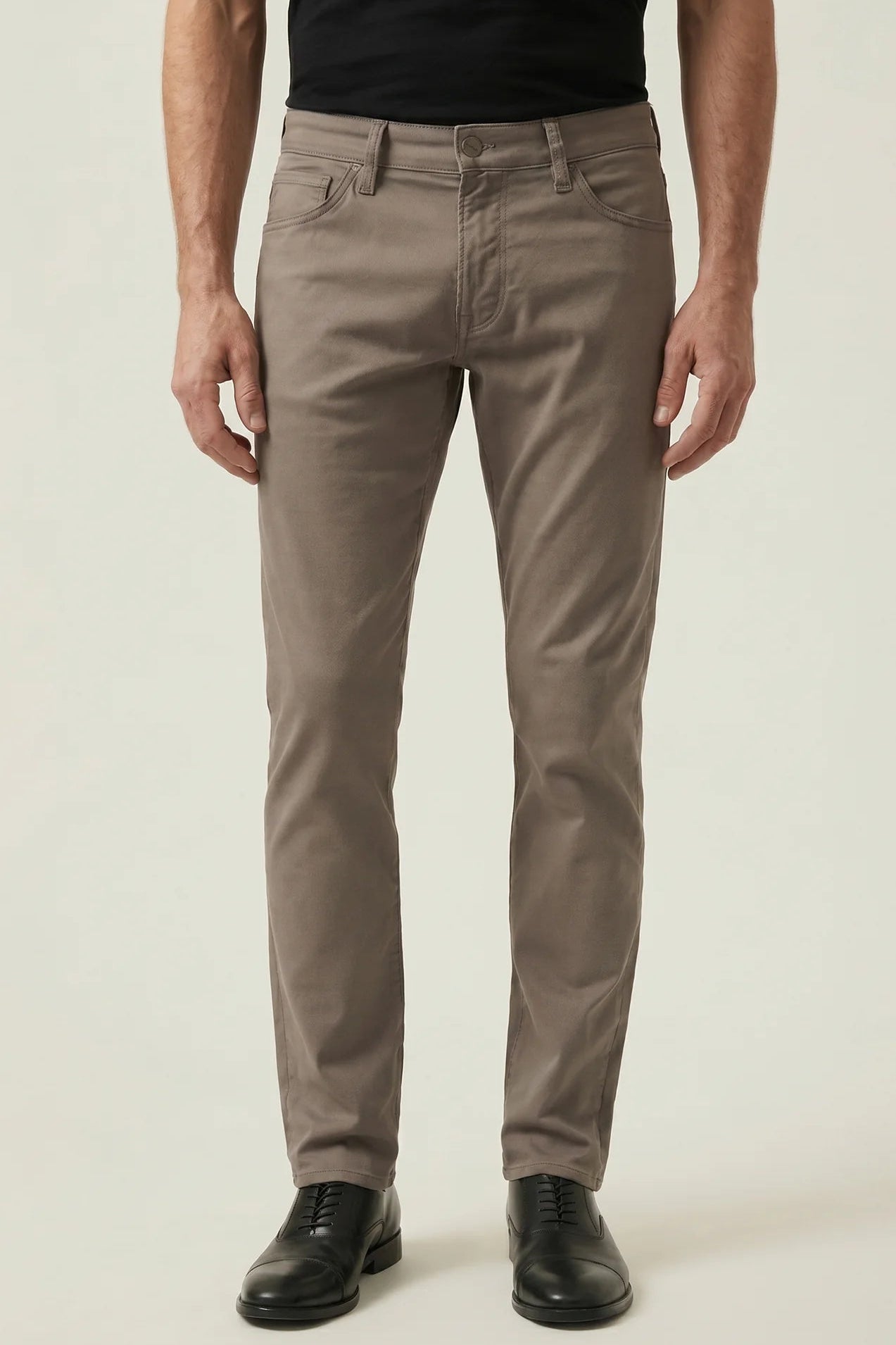 A man wearing slim-fit taupe brown trousers, a black t-shirt, and black leather dress shoes, standing against a light beige studio background.