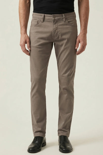 A man wearing slim-fit taupe brown trousers, a black t-shirt, and black leather dress shoes, standing against a light beige studio background.
