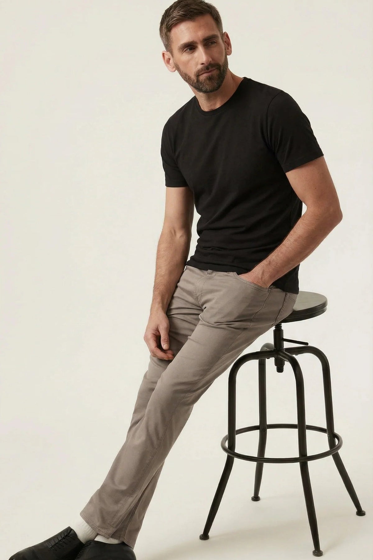 Man sitting on a stool wearing a black t-shirt, beige pants, and black shoes on a white background