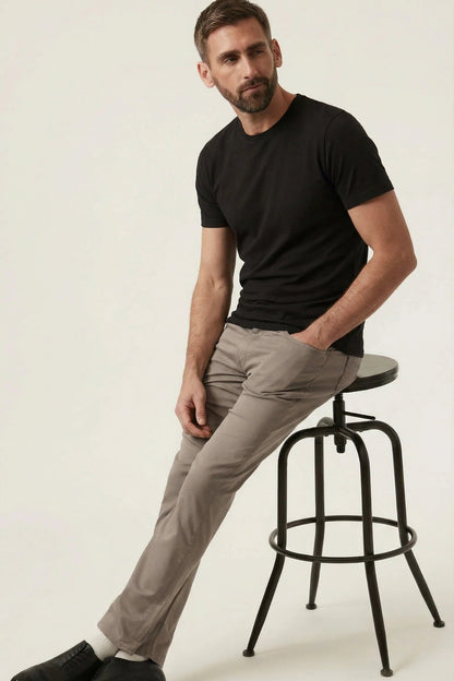 Man sitting on a stool wearing a black t-shirt, beige pants, and black shoes on a white background