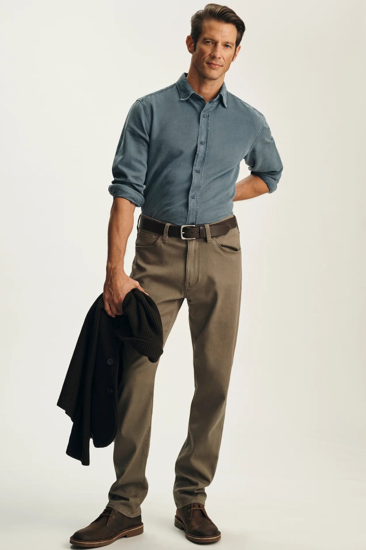 A man modeling a blue button-down shirt, brown trousers, and brown shoes, holding a black jacket in one hand against a light gray studio background.