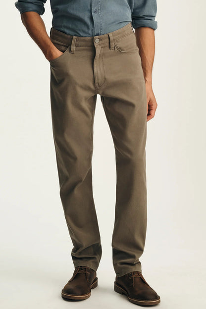 A man wearing slim-fit brown trousers, a blue button-down shirt, and brown leather shoes, standing against a plain light background.