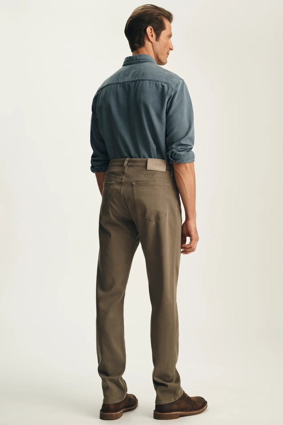 A man shown from the back wearing a slate blue button-down shirt, olive brown trousers, and brown shoes, standing against a light gray studio background.