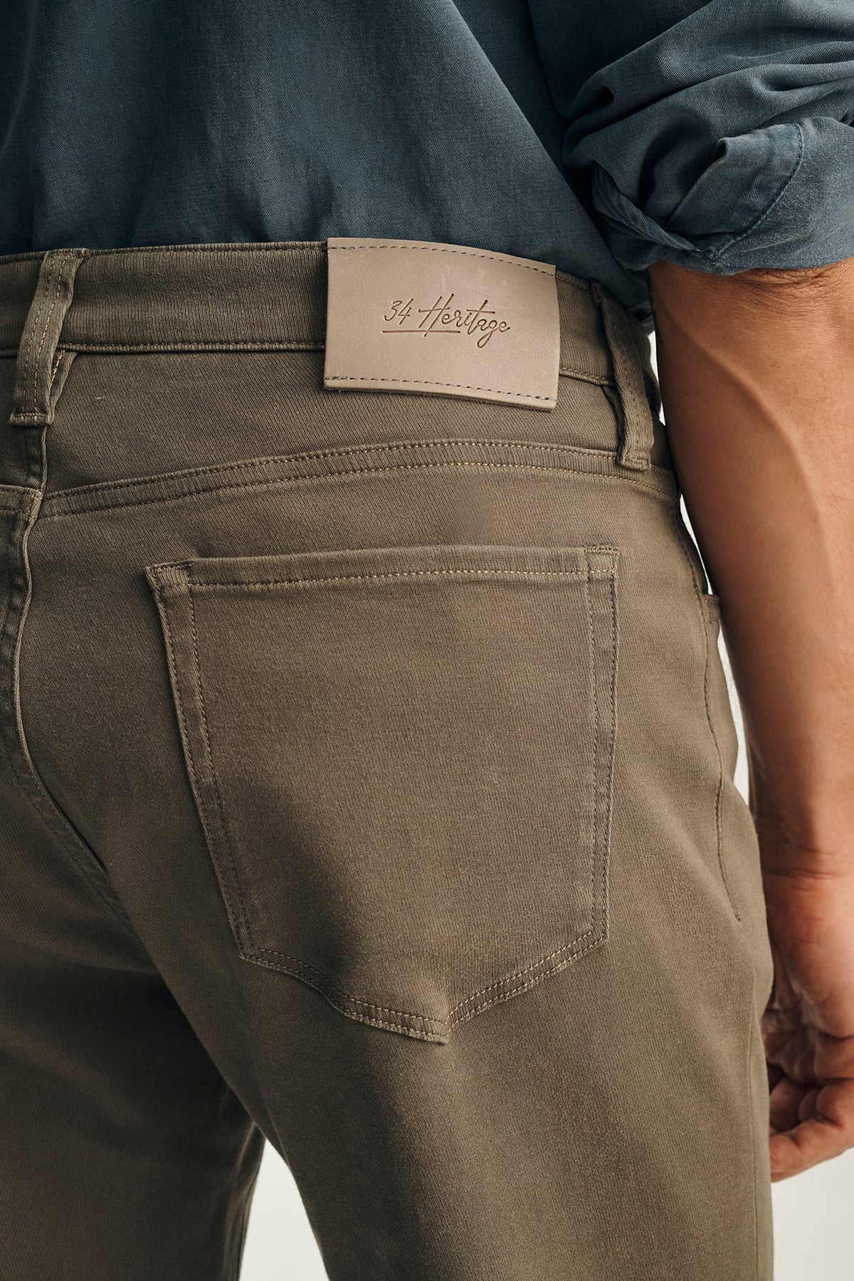 Close-up back view of a person wearing olive green denim jeans with a tan leather St. Harengo brand patch at the waistband, paired with a rolled-up blue shirt.