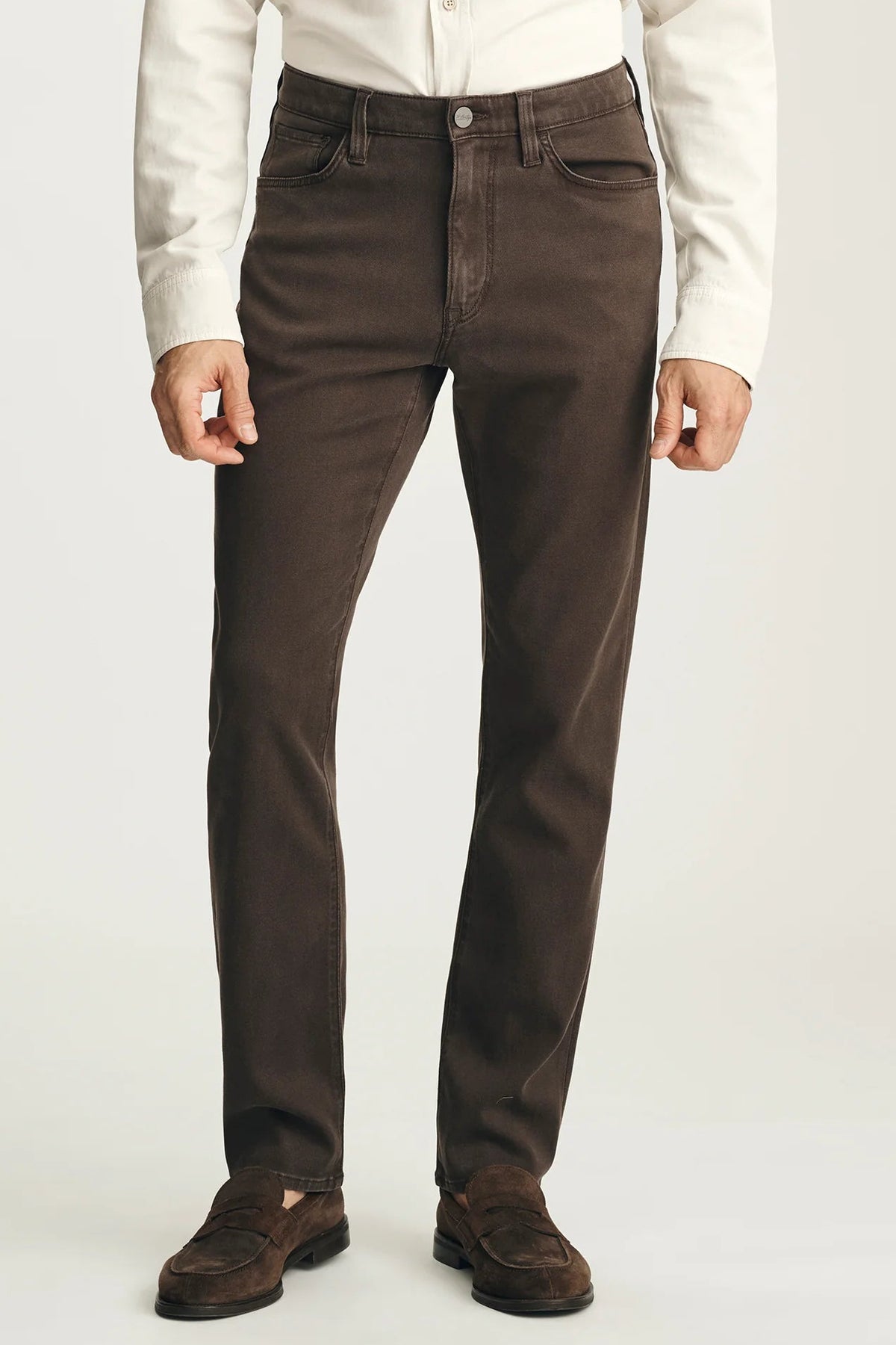 A person wearing dark brown slim-fit jeans, a white long-sleeve shirt, and brown suede loafers, shown from the waist down against a light gray studio background.
