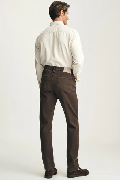 A man shown from the back wearing a white button-down shirt, dark brown trousers, and dark brown loafers, standing against a light gray studio background.