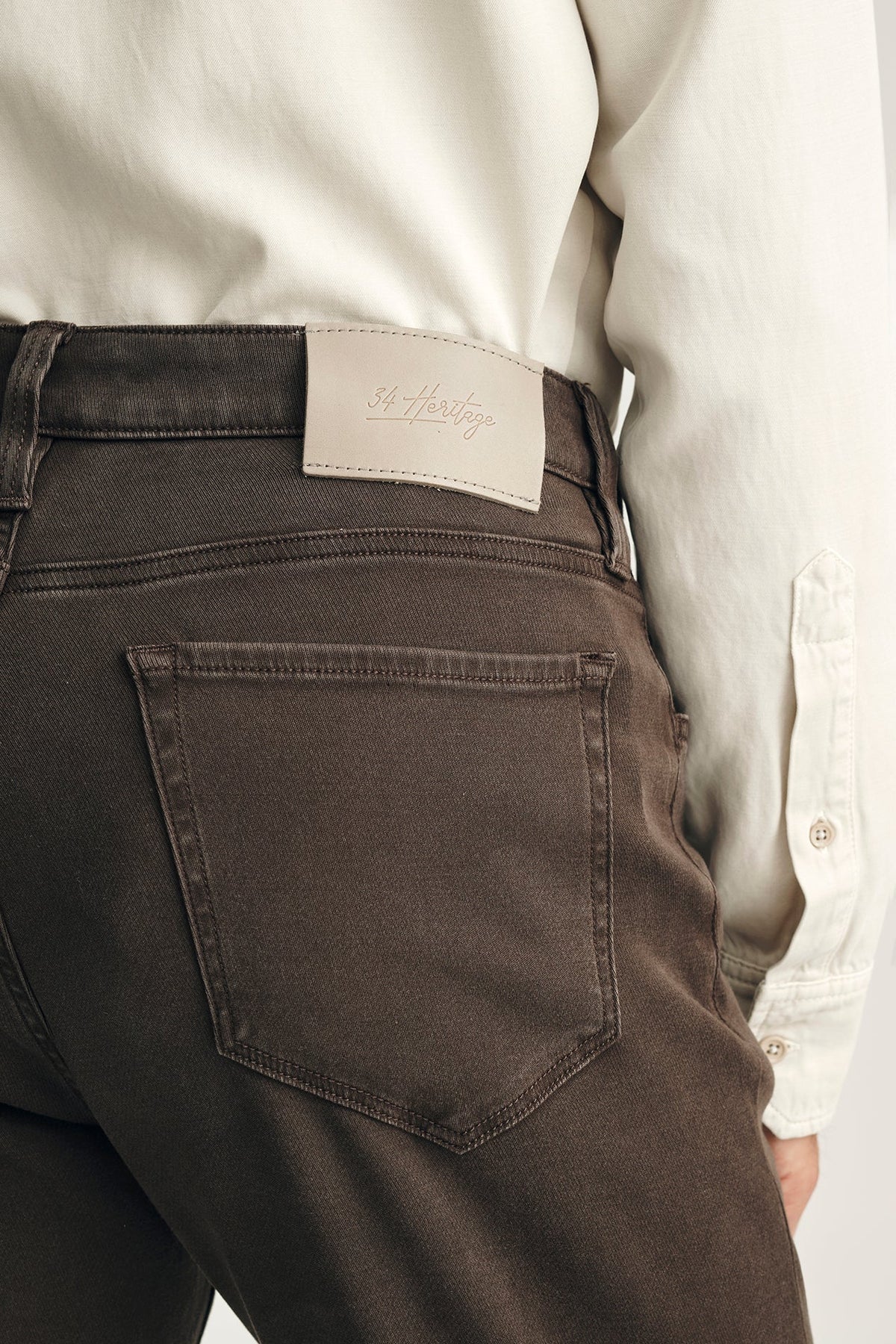 Close-up of the back waist and back pocket of dark brown jeans worn by a model, paired with a cream-colored long-sleeve shirt. A light beige leather patch with the brand name 'St.
