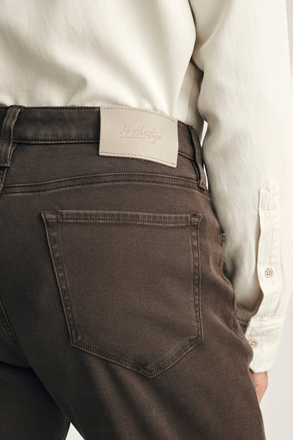 Close-up of the back waist and back pocket of dark brown jeans worn by a model, paired with a cream-colored long-sleeve shirt. A light beige leather patch with the brand name 'St.