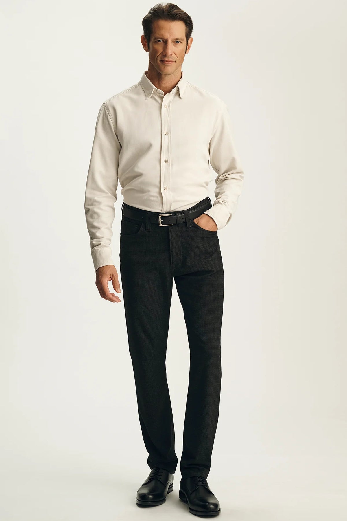 A man modeling a long-sleeve cream button-down shirt tucked into black dress pants with a black belt and black dress shoes, standing against a plain light gray studio background.