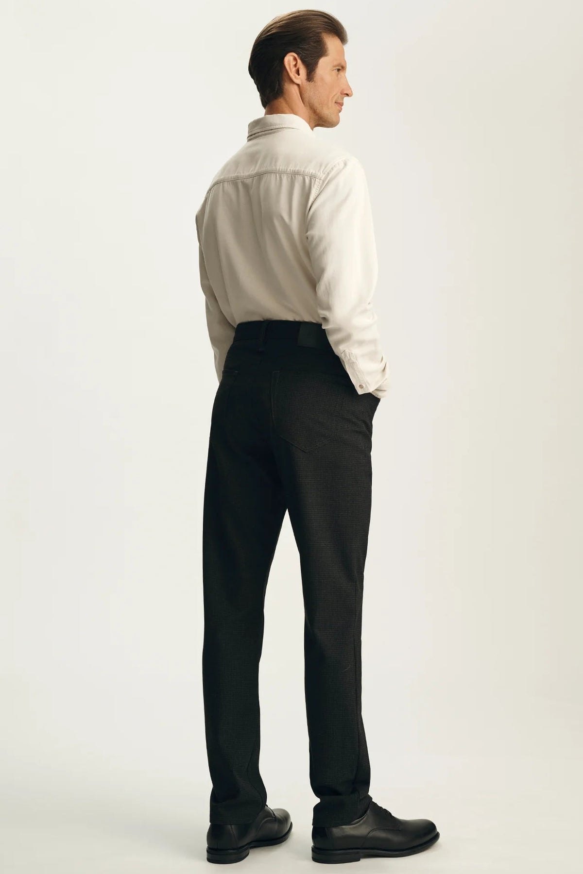 A man shown from the back wearing a light beige button-down shirt tucked into black dress pants and black shoes, standing against a plain light gray studio background.