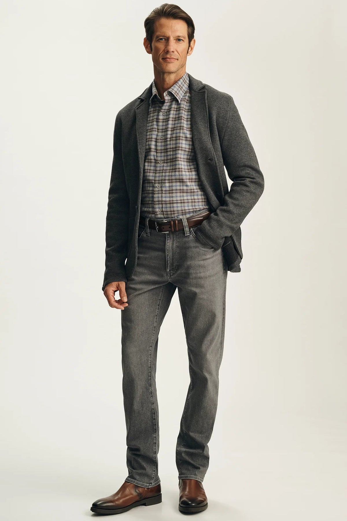 A man modeling a gray blazer, gray plaid shirt, gray jeans, and brown leather dress shoes against a light gray studio background.