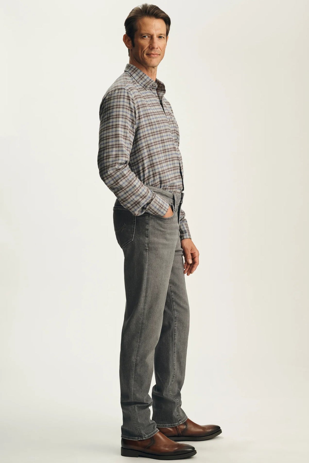 A man wearing a brown and gray plaid button-down shirt, gray jeans, and brown leather shoes, standing in profile against a light gray studio background.