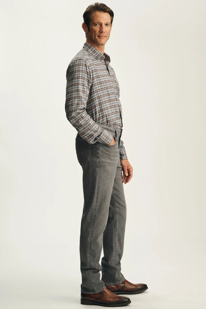A man wearing a brown and gray plaid button-down shirt, gray jeans, and brown leather shoes, standing in profile against a light gray studio background.