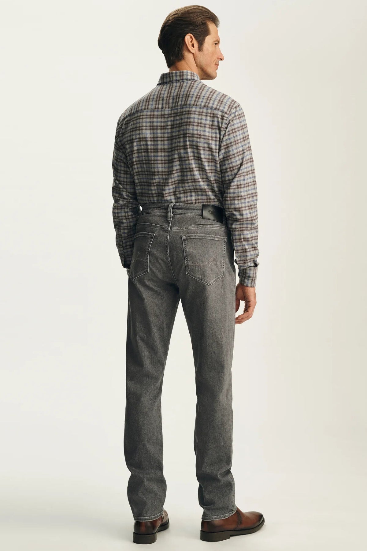 A man shown from the back wearing a gray plaid button-down shirt tucked into gray jeans with a black belt, paired with brown leather shoes, against a light gray studio background.