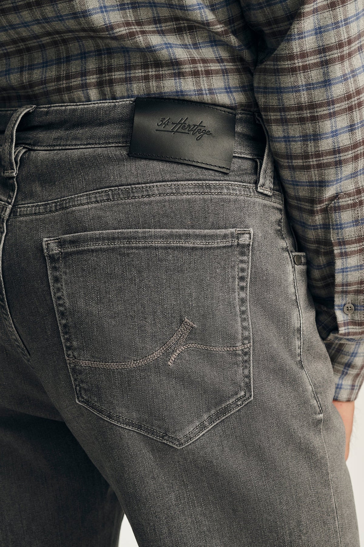 Close-up of the back waistband of dark gray washed denim jeans, showing a black leather patch with the 'Softer' brand logo, worn with a plaid shirt.
