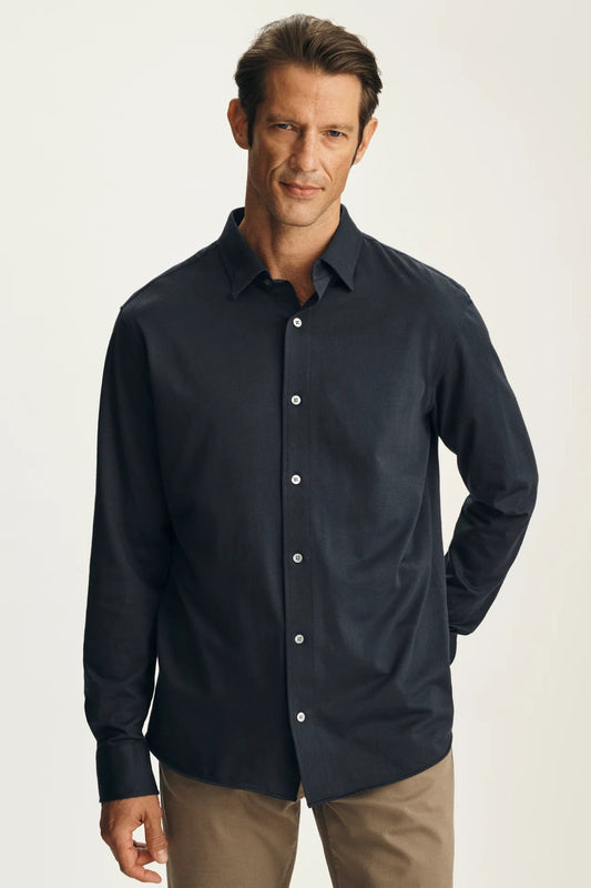A man wearing a dark navy blue long-sleeve button-down shirt and khaki pants, standing against a light beige studio background.