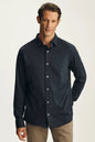 A man wearing a dark navy blue long-sleeve button-down shirt and khaki pants, standing against a light beige studio background.