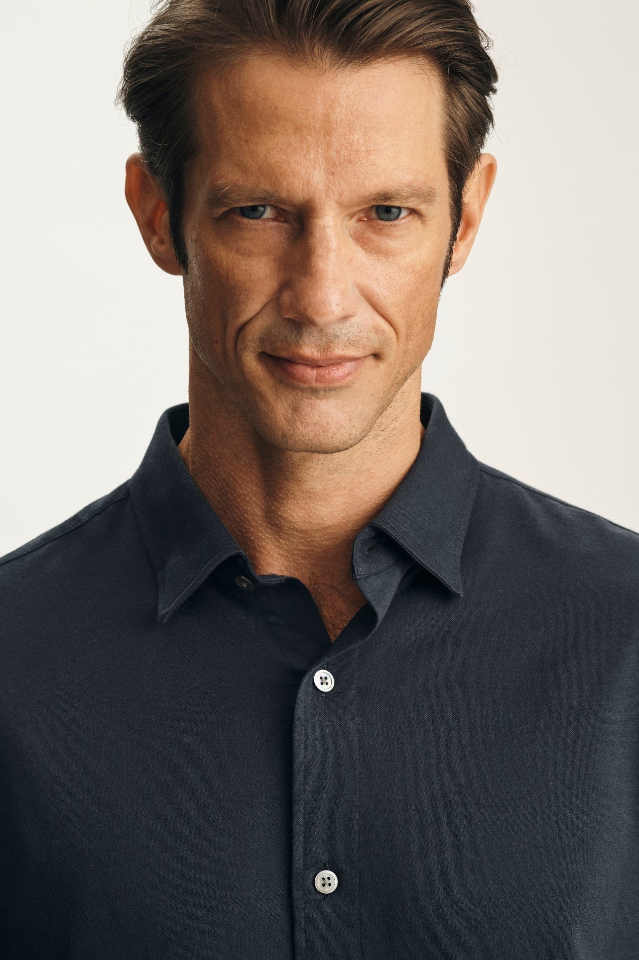 A close-up portrait of a man wearing a dark navy blue button-down shirt against a light off-white background.